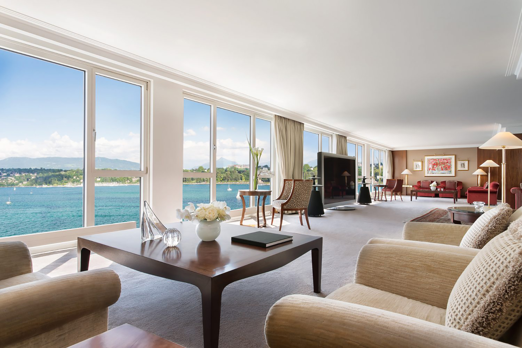  A living room with a large glass window looking out onto a lake and mountains with a coffee table, chairs, and sofa in the foreground.